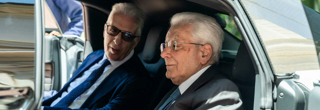 Sergio Mattarella e un altro uomo in abito blu salgono a bordo di un'auto grigia.