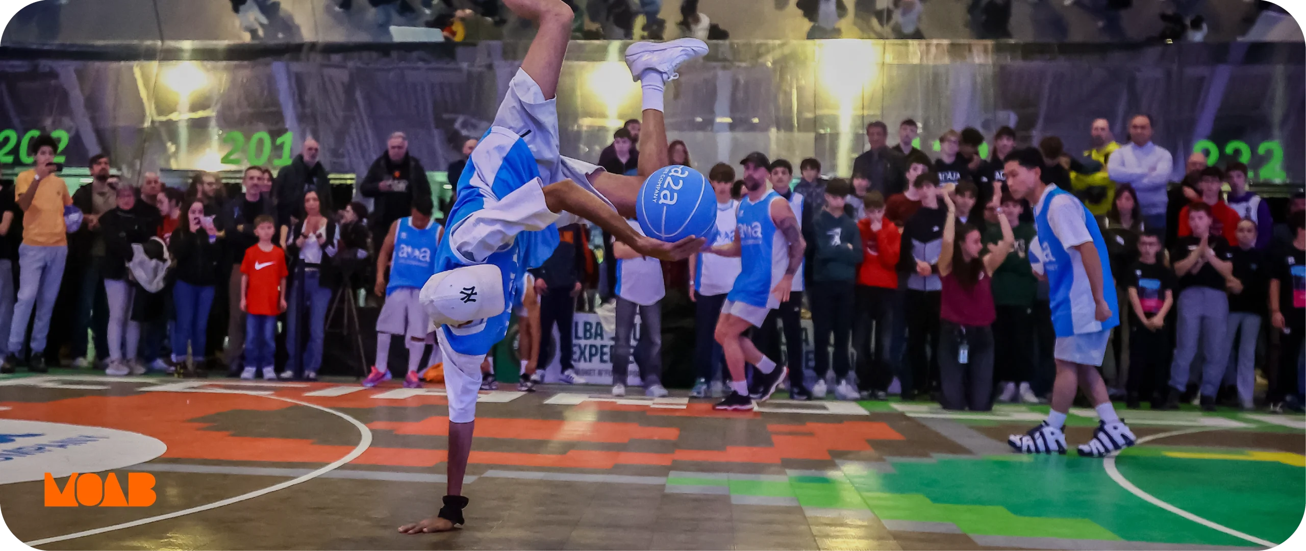 Banner agenzia marketing sport: atleta esegue una verticale con palla da basket, pubblico sullo sfondo. Spettacolo sportivo.