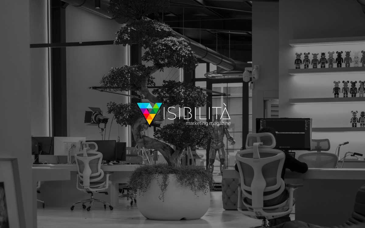 Logo "Visibilità marketing magazine" con triangolo multicolore su bonsai in ufficio moderno in bianco e nero.